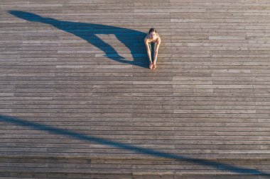 Tahta zeminde oturup kameraya bakarken, kollarını bacakları boyunca uzatan ve esneklik gösteren tanınmayacak kadar sağlıklı bir kadının üst görüntüsü.