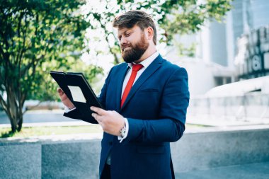 Ciddi sakallı iş adamı takım elbise giymiş ofis binalarının yanında duruyor. Yemek molasında toplantı için hazırlanan klasördeki belgeleri okuyor.
