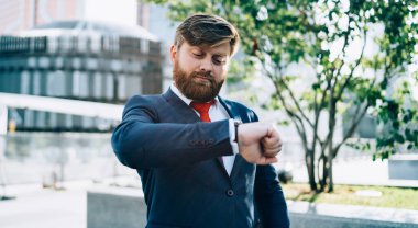 İş merkezine karşı caddede duran kırmızı kravatlı ciddi sakallı iş adamı öğle arasında kol saatine bakıyor.