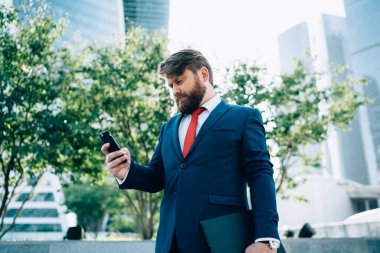 Takım elbiseli ve kırmızı kravatlı konsantre sakallı işadamı iş merkezinin yanındaki klasörde duran akıllı telefondan mesaj okuyor.