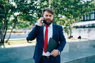 Endişeli sakallı, şık takım elbiseli bir işadamı iş merkezinin yanında elinde dosyayla kameraya bakarak telefonda konuşuyor.