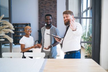 Kendine güvenen sakallı, resmi kıyafetli, neşeli etnik çifte mutfağını gösteren tombul bir adam modern şehir dairesinde duruyor.
