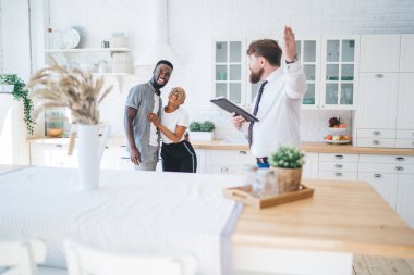 Profesyonel sakallı, resmi kıyafetli, yakışıklı bir adam. İlgili etnik çifte mutfağı gösteriyor. Modern, güneşli bir dairede masanın yanında duruyor.