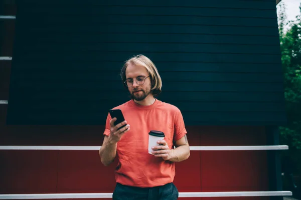 Young bearded modern guy with long hair in red t-shirt texting on mobile phone holding cup of takeaway coffee in city street