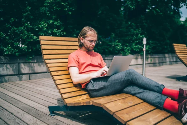 Young red bearded man in glasses with long hair in red t shirt red socks working with laptop laying on lounge