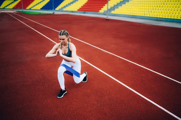 Spor kıyafetleri ve spor ayakkabıları giyen konsantre sarışın kadın stadyumdaki antrenman sırasında lastik kurdeleyle egzersiz yapıyor ve göğsüne yakın elle saldırıyor. 
