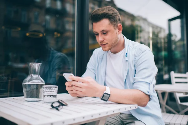 若い知的男性身に着けている青シャツ白Tシャツと手首の腕時計メッセージングオン携帯電話でテーブルに座って屋外テラス — ストック写真