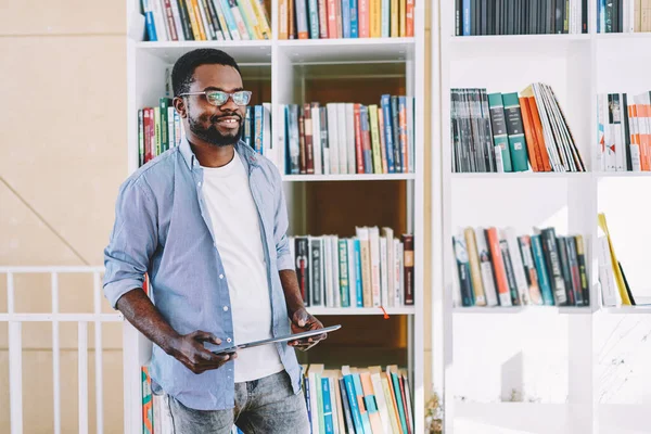 Smart African American man in glasses standing nearby bookshelves and holding tablet for studying and searching information in light library