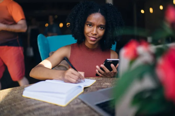 在咖啡店准备考试的时候 穿着休闲装 面带笑容的黑人女士在笔记本浏览手机上写便条 — 图库照片