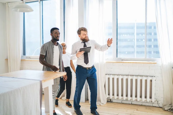 Convincing bearded chubby man in formal outfit with clipboard showing kitchen to interested ethnic couple while walking along modern sunny apartment