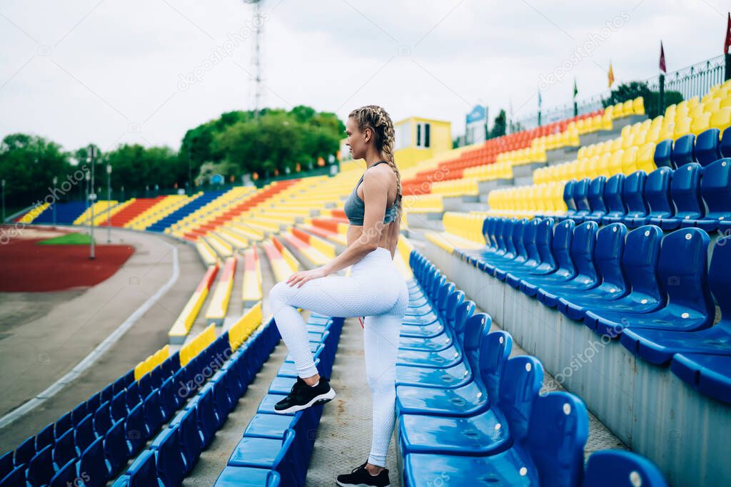 Side view of athletic blond female with braids and in sportswear standing with left leg on seat of empty stadium and looking forward