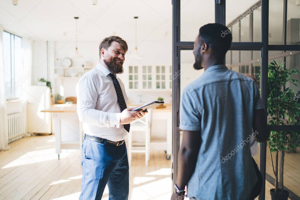 Laughing bearded adult formal man with tablet meeting diverse clients and chatting in hall of modern light apartment standing opposite