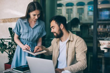 Sakallı etnik adam, sokak kafesindeki kadın çalışanla serbest çalışma projesini tartışırken not defterindeki verilere gülümsüyor ve onları işaret ediyor.