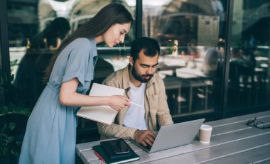 Not defteri ve kalemi olan genç bir kadın kafeterya masasının yanında duruyor ve birlikte serbest çalışırken dizüstü bilgisayar kullanan sakallı etnik adamı denetliyor.