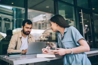 Akıllı telefonlu genç bir kadın, sokak restoranındaki masada otururken bilgisayarlı etnik erkek arkadaşının çalışmalarını planlayıp kontrol ediyor.