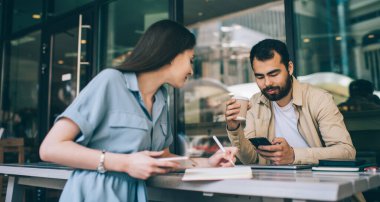 Not defterine yazan genç bir kadın akıllı telefonlu ve içki içen erkek iş arkadaşına bakıyor.