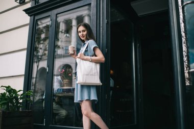 Modern, yoğun bir bayanın gündelik kıyafetlerini omuzlarında modaya uygun beyaz bir bez torbayla tek kullanımlık kahve bardağını tutarken ve kahve dükkanından çıkarken kameraya bakarken... 