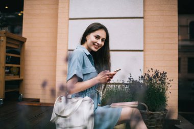 Şehir binasının önünde oturmuş akıllı telefondan mesajlaşırken gülümseyen ve kameraya bakan mutlu genç öğrencinin yan görüntüsü. 