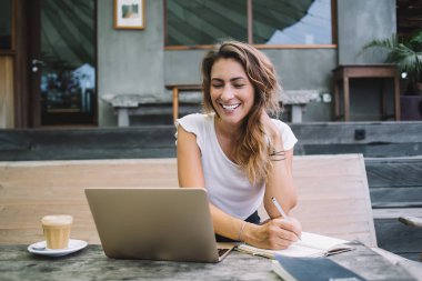 Neşe dolu güzel bir kadın deftere yazıp laptopa bakarken bir yandan da kafeteryada elinde kahve fincanıyla oturuyor.