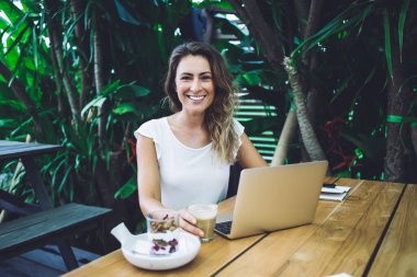 Mutlu bayan elinde bir fincan taze kahve tutarken ve kameraya gülümserken açık kafedeki yeşil bitkilerin yanındaki masada modern dizüstü bilgisayarlara bakarken.
