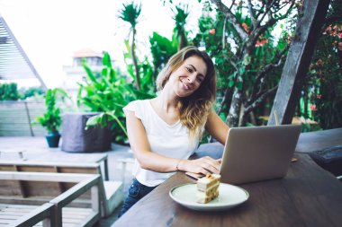 Açık hava restoranındaki tatlı masasının yanında otururken gündelik kıyafetleriyle gülümseyen ve modern laptopları karıştıran mutlu yetişkin bir kadın.