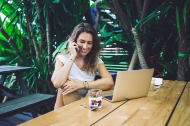 Arkadaşça gülümseyen iyimser kadın bahçedeki yeşil bitkilerin yanında tatlı ve dizüstü bilgisayarla kafeterya masasında otururken akıllı telefondan konuşuyor.