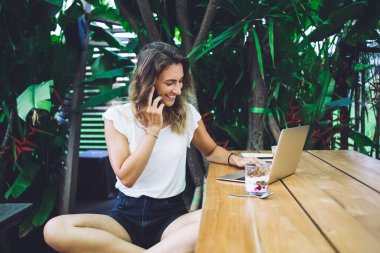 Zarif kıyafetli iyimser bir bayan gülümseyip laptopunu kullanırken masada tatlıyla oturuyor ve telefonlarına cevap veriyor. 