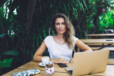 Beyaz gömlek giyen genç bir kadının laptop ve tatlıyla kahve molası verirken kafenin ahşap masasında oturup tropik bitkilerle süslenmiş bir şekilde kameraya bakması.