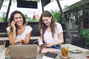 Neşeli genç kız öğrenciler not defterlerinde notlar okuyor ve açık kafede oturmuş birlikte ders çalışırken akıllı telefon konuşmaları yapıyorlar.