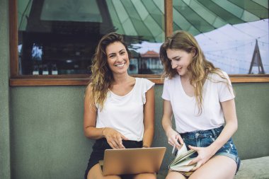 Mutlu kız öğrenciler not defterini açıp dizüstü bilgisayar kullanırken binanın dışında büyük pencereli bir bankta oturuyorlar ve birlikte ödev yapıyorlar.