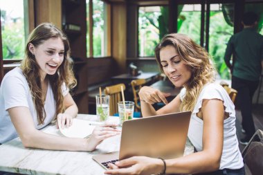 Neşeli genç bayan dizüstü bilgisayarı işaret ediyor ve hem rahat bir kafede oturan hem de projeyi tartışan şaşkın bir bayan arkadaşa bilgi veriyor.