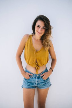 Keyifli, rahat, rahat, rahat, modaya uyan genç bir kadın üstte ve güneşli bir günde beyaz duvara yaslanan şortlar.