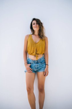 İçerik, tasasız, rahat, modaya uyan genç bir kadın üstte ve güneşli bir günde beyaz duvara yaslanan şortlar.