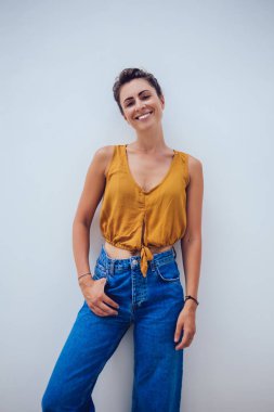 Kot pantolon ve turuncu gömlekli şık bir kadın. Elleri cebinde gülümsüyor ve kameraya bakıyor.