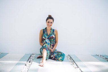 Maksimum gösterişli elbiseli mutlu bir kadın bacak bacak üstüne atarak rahatlarken duvarda poz veriyor ve beyaz arka planda kameraya bakıyor.