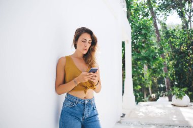 Kıvırcık düşünceli kadın, yeşil ışık bahçesindeki beyaz duvarlara yaslanmış cep telefonuyla sörf yaparken konsantre olmuş.