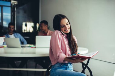 Neşeli kadın analitik uzmanı cep telefonuyla telefon görüşmesi yapan, arka planda erkek ortaklarla ofisin içinde oturan müşteriyle hücresel istişare sırasında kağıt ders kitabından bilgi kontrol ediyor.