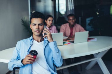 Ofiste elinde bir fincan kahve tutan etnik bir adam ve çok ırklı meslektaşlarıyla otururken akıllı telefonlara danışmanlık yapıyor.