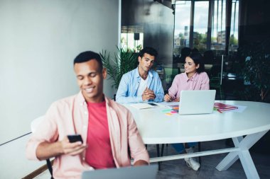 Ofiste dizüstü bilgisayarla çalışan ve sosyal medyada akıllı telefondan mesaj okuyan gülümseyen etnik bir adam. 