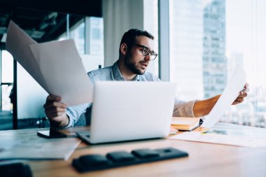 Finansal bilgileri ve verileri kontrol etmek için elinde evrak tutan genç adam modern netbook ile masada oturuyor ve muhasebe üzerine yoğunlaşıyor, akıllı erkek tüccar belgelerden rapor analiz ediyor.