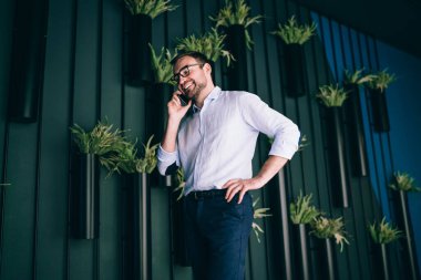 Modern ofisteki saksı bitkileriyle dekore edilmiş duvarın yanında dururken akıllı telefondan gülümseyen ve konuşan mutlu erkek yöneticinin alçak açısı.