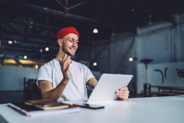 Kırmızı bereli neşeli ve hevesli adam modern geniş ofisteki masada Stylus 'la çalışırken tablet üzerinde video görüşmesi yapıyor. 