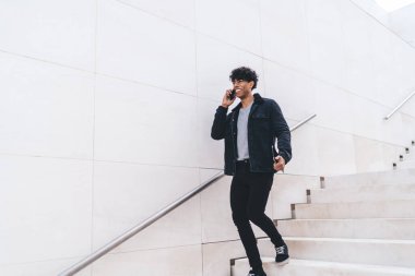 Modern stil, mutlu, gülümseyen, Afro-Amerikalı bir adam. Günlük kıyafetleriyle telefonda konuşurken gün ışığında şehir merdivenlerinde yürürken.