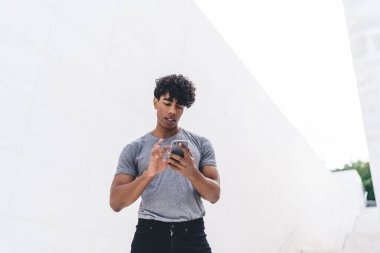 Gri tişörtlü, yakışıklı, kıvırcık, yetişkin bir erkek beyaz duvarın önünde caddede sörf yapıyor.  