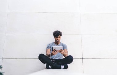 Şok olmuş genç, sıradan kıyafetlerle yakışıklı bir erkek bacak bacak bacak üstüne atıyor ve dışarıda kulaklıklarla video seyrediyor.