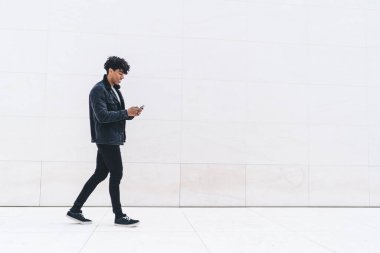 Gülümseyen, genç, kıvırcık, siyah ceketli, kot pantolonlu, beyaz mermer duvarın önünde tek başına yürürken yakışıklı bir adam. 