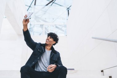 Parlak odadaki beyaz merdivenlerde oturan şık kot elbiseli neşeli kıvırcık saçlı Amerikalı adam selfie çekiyor ve gülümsüyor.