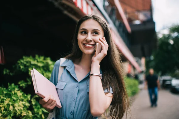 快乐的年轻女性 一边笑着一边望着远方 一边用智能手机说话 走在城市街道模糊的背景上 — 图库照片