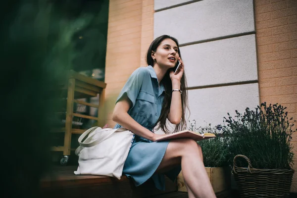 膝の上に開いたノートを持ち 街の通りの外のビルのベンチに座っている間にスマートフォンで話している青いドレスのスタイリッシュな若い女性 — ストック写真