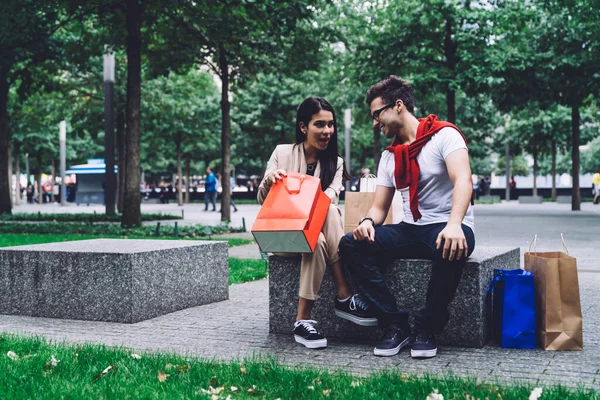 戴眼镜的男朋友和穿着时髦服装的女朋友在公园外面模糊的背景下购物后坐在石凳上 — 图库照片